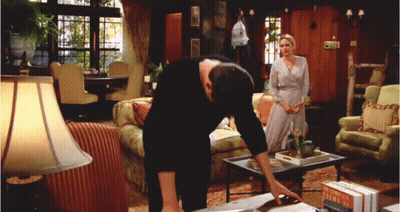 A Man Leans Over A Table In A Living Room With A Woman Standing In The Background GIF