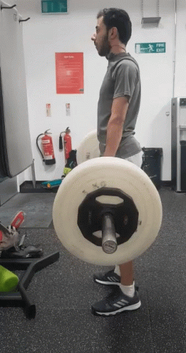 A Man Lifting A Barbell In A Gym With A Fire Extinguisher On The Wall Behind Him GIF