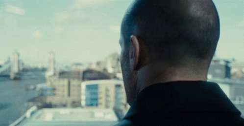 A Man Looking Out A Window With Buildings In The Background GIF