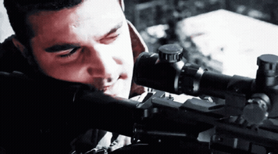 A Man Looking Through A Sniper Scope With A Red Light Behind Him GIF