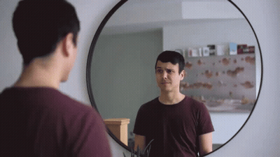 a man looks at himself in a round mirror with a reflection of a shelf with books on it