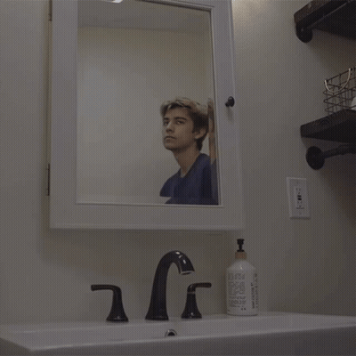 A Man Looks At His Reflection In A Mirror In A Bathroom GIF