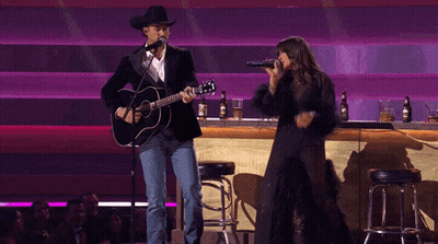 A Man Playing A Guitar And A Woman Singing Into A Microphone On A Stage GIF