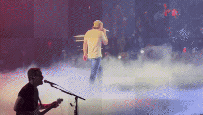 A Man Playing A Guitar And Singing Into A Microphone On A Stage GIF