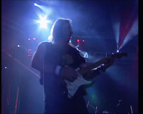 A Man Playing A Guitar On A Stage With A Blue Light Behind Him GIF
