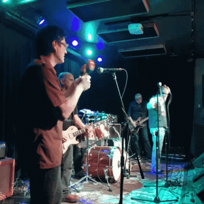 A Man Playing A Guitar Stands In Front Of A Microphone GIF