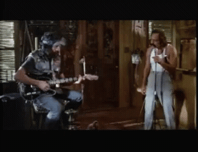 A Man Playing A Guitar While Another Man Sings Into A Microphone GIF