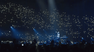 A Man Playing A Keyboard On A Stage With A Crowd Behind Him GIF