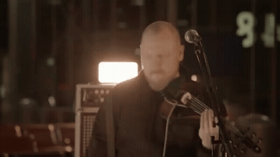 A Man Playing A Violin In Front Of A Microphone With The Number 9 On The Wall Behind Him GIF