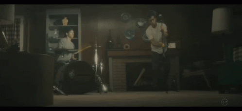 A Man Playing Drums In A Living Room With A Man Playing A Guitar In The Background GIF