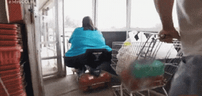 A Man Pushes A Shopping Cart With A Woman In A Wheelchair In The Background GIF