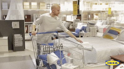 A Man Pushing A Shopping Cart In An Ikea Store With A Bed In The Background GIF