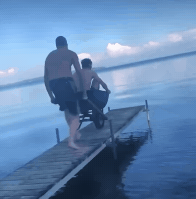 A Man Pushing Another Man In A Wheelbarrow Down A Pier GIF