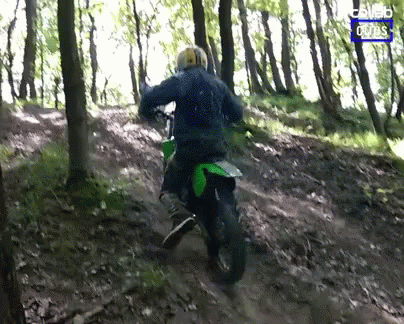 A Man Riding A Dirt Bike In The Woods With A Blue Sign That Says Combo Rides GIF