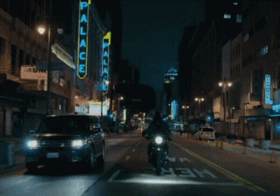 A Man Riding A Motorcycle Down A City Street With A Neon Sign That Says Palace GIF