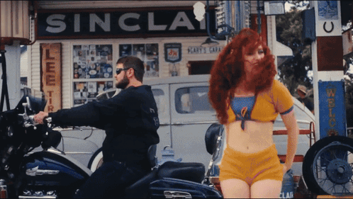 A Man Riding A Motorcycle Next To A Woman In Front Of A Sinclair Sign GIF