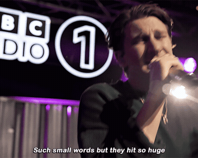 A Man Singing Into A Microphone In Front Of A Bbc Logo GIF