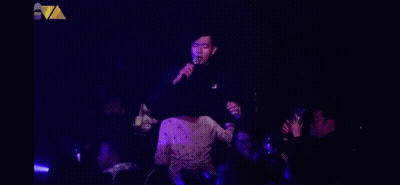 A Man Singing Into A Microphone In Front Of A Crowd In A Dark Room GIF
