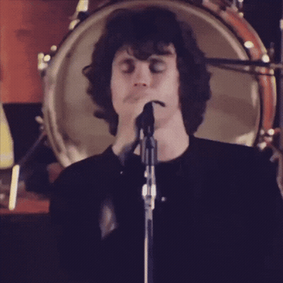 A Man Singing Into A Microphone In Front Of A Drum Set GIF