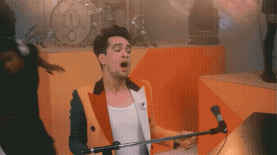 A Man Singing Into A Microphone In Front Of A Drum Set With An Exclamation Point On It GIF