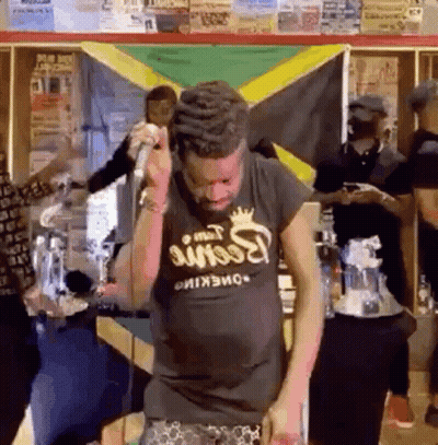 A Man Singing Into A Microphone In Front Of A Jamaican Flag GIF
