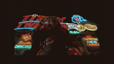 A Man Singing Into A Microphone In Front Of A Lit Up Sign That Says Titty Twist GIF