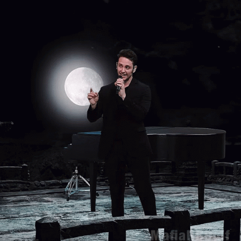 A Man Singing Into A Microphone In Front Of A Piano GIF