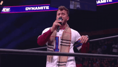 A Man Singing Into A Microphone In Front Of A Sign That Says Dynamite On It GIF