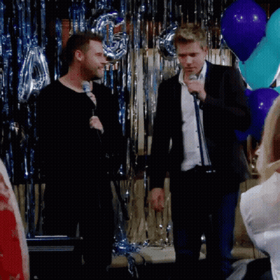 A Man Singing Into A Microphone In Front Of Balloons And Tinsel GIF