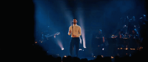 A Man Singing Into A Microphone On A Stage In Front Of A Crowd GIF