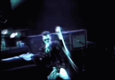 A Man Singing Into A Microphone On A Stage With A Light Behind Him GIF