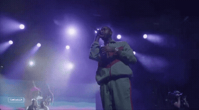 A Man Singing Into A Microphone On A Stage With Purple Lights Behind Him And A Sign That Says Campella GIF
