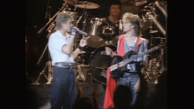 A Man Singing Into A Microphone While Another Man Plays A Guitar On Stage . GIF