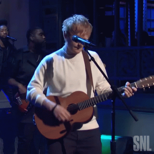 A Man Singing Into A Microphone While Holding A Guitar With The Snl Logo In The Background GIF