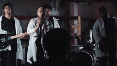 A Man Singing Into A Microphone While Playing A Guitar And A Drum Set GIF