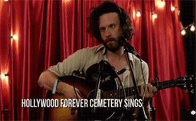 A Man Singing Into A Microphone While Playing A Guitar With The Words Hollywood Forever Cemetery Sings Written Below Him GIF