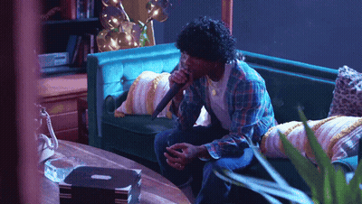 A Man Singing Into A Microphone While Sitting On A Green Couch GIF