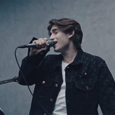 A Man Singing Into A Microphone While Wearing A Black Jacket With The Letter C On It GIF