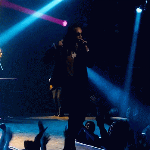A Man Singing Into A Microphone With A Crowd Behind Him GIF