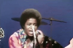 A Man Singing Into A Microphone With A Drum Set Behind Him GIF