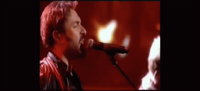A Man Singing Into A Microphone With A Guitar In The Background GIF