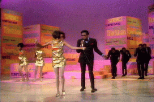 A Man Singing Into A Microphone With A Woman Dancing In Front Of A Stack Of Boxes That Say Temptations GIF