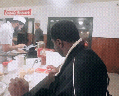 A Man Sits At A Counter In Front Of A Sign That Says " Quality Forever " GIF