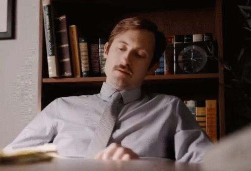A Man Sits At A Desk In Front Of A Bookshelf With A World Book Atlas On It GIF