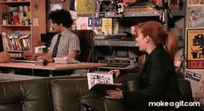 A Man Sits At A Desk While A Woman Sits On A Couch With A Book In Her Hand GIF