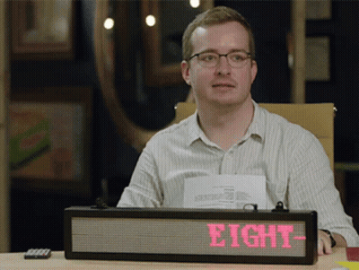 A Man Sits At A Desk With A Sign That Says Eight On It GIF