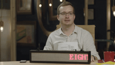 A Man Sits At A Desk With A Sign That Says Eight On It GIF