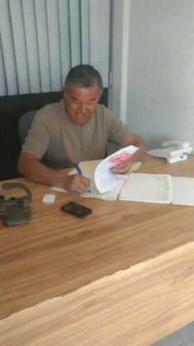 A Man Sits At A Desk Writing On A Piece Of Paper With A Pen GIF