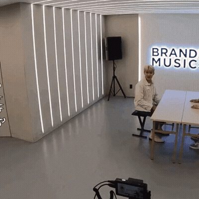 A Man Sits At A Table In A Room With A Sign That Says Brand Music GIF