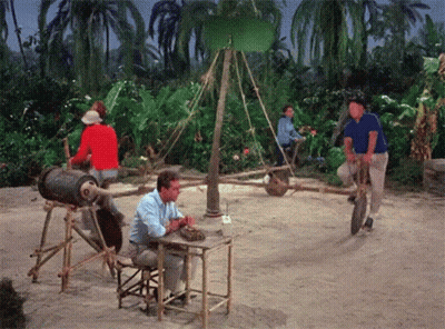 A Man Sits At A Table While Another Man Rides A Wheelbarrow GIF